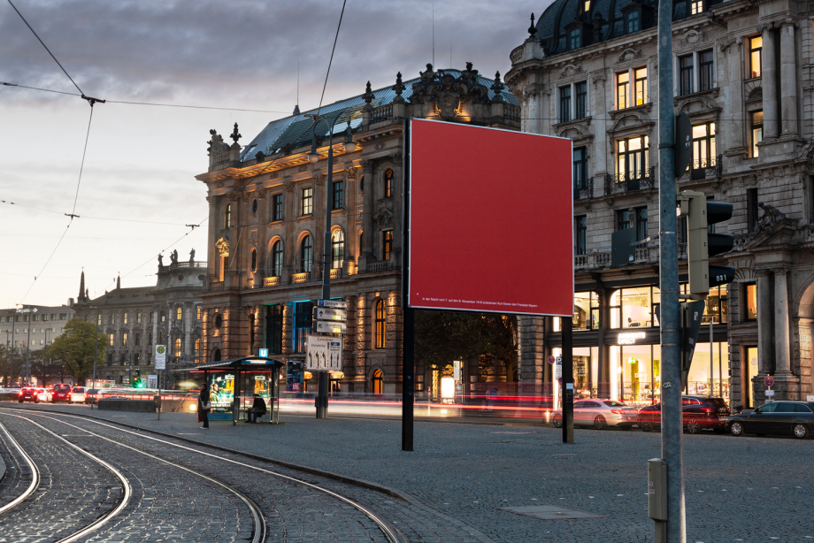 Diagonal view of the billboard at Lenbachplatz in the evening rush hour. The motif is designed in bright red monochrome with a footnote on Kurt Eisner in white lettering.